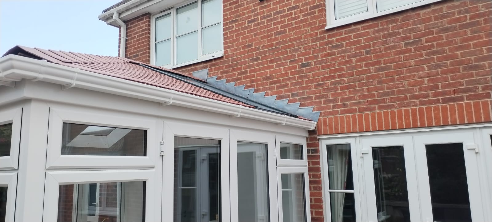 White uPVC conservatory windows and French doors alongside new casement windows on a red-brick detached home with a tiled conservatory roof and lead flashing